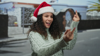 Woman wearing santa hat gestures on urban street dressed in sweater, capturing a candid moment outdoors with expressive attitude and festive vibes. © Krakenimages.com