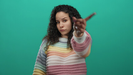Woman gesturing no with hand towards camera against isolated green background, wearing colorful...