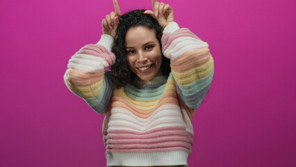 Hispanic woman wearing colorful sweater playfully making horn gesture against bright pink background