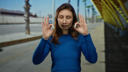Woman meditating outdoors in city street showcases calm expression with closed eyes, wearing blue...
