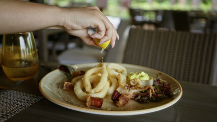 Woman squeezing lemon over calamari on a plate in a restaurant setting with a drink on the table, capturing an appetizing dining experience.
