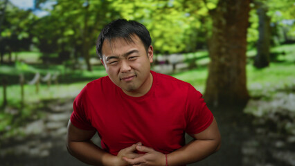 Fototapeta premium Man in red shirt clutches stomach painfully in a sunny park surrounded by lush greenery, highlighting discomfort outdoors.