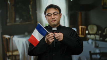 Man holding french flag in a restaurant setting, wearing clerical attire with a cross visible on a...