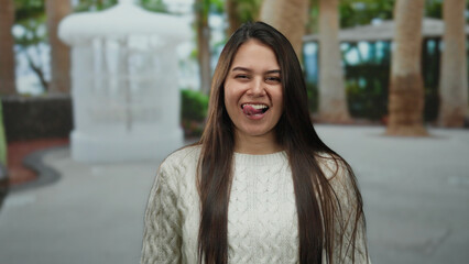 Fototapeta premium Woman in white sweater playfully sticking tongue out in outdoor park setting with palm trees and structures in the background.