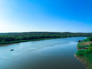 A wide tranquil river flows calmly under a clear blue sky, surrounded by lush green trees and forest-covered hills. 