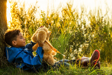 In the enchanting embrace of golden hour, a little boy sits on the grass beneath the sprawling...