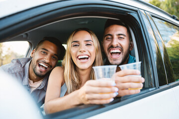 Happy friends in a car, smiling and raising drinks. Joyful road trip or party moment with young adults looking at the camera and cheering from the window.