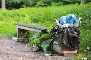 An Overflowing Public Trash Can With Plastic Bags And Litter Spilling Out, Next To A Wooden Bench...