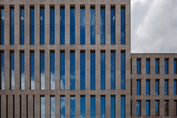 A close-up view of a contemporary building facade featuring a grid of tall, narrow blue windows reflecting the sky, set against light brown stone cladding.