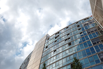A low-angle view of a contemporary building facade dominated by a grid of reflective glass windows, some slightly ajar, reflecting a cloudy sky. 
