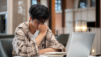 Man in plaid shirt holding or massaging his eye in front of a laptop and book sitting at cafe table.
