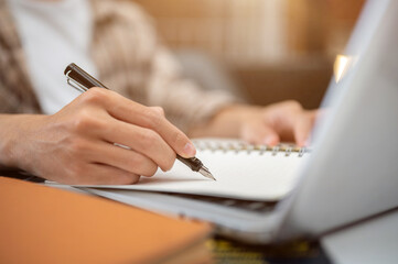 Close up of a man hand holding fountain pen writing in notebook over laptop sitting at table in cafe
