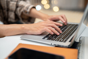 Close up of a man's both hands typing on laptop with phone atop book sitting at white table in cafe.