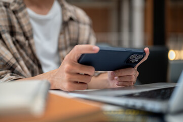 Close up of a man hands holding phone watching video playing game near laptop and book on cafe table