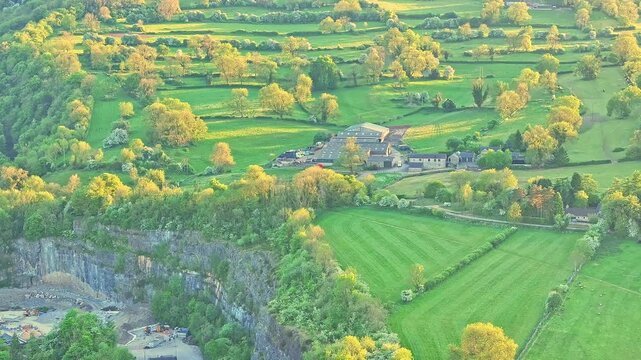 Aerial view captures farmlands and outbuildings nestled among patterned fields and trees, with a dramatic limestone cliff nearby, Matlock Sparrow Park area, Derbyshire, rural English countryside scene