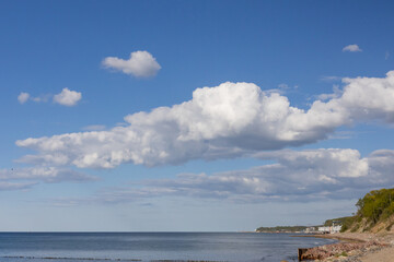 Wide coastal view with calm sea, rocky shore, and cumulus clouds under clear blue sky.