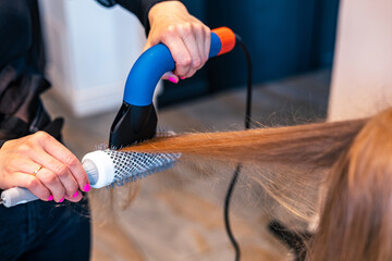 Female hairstylist drying and straightening long blonde client hair with comb and hairdryer in...