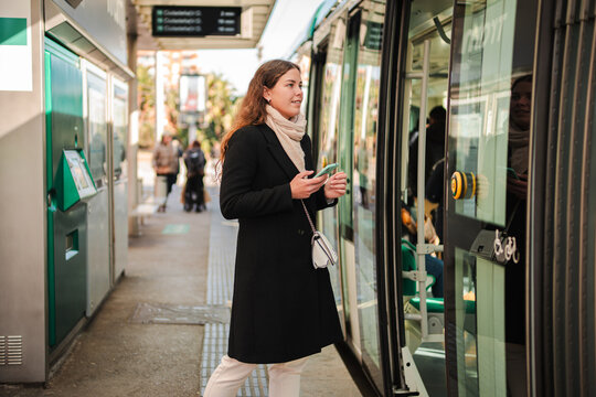 A female passenger waiting at the subway station entrance, looking at her phone while eagerly anticipating the arrival of her transport, ready to embark on her next adventure in the busy city life.