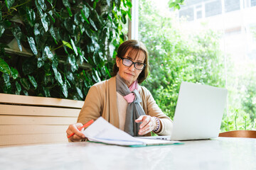 Mature woman working at a cafe with a laptop and financial documents. She reviews papers intently in a green, cozy setting, symbolizing focus, professionalism, and modern entrepreneurship.