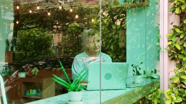 Thoughtful cafe owner works on a laptop while gazing out the window in a lush, plant-filled shop. Scene captures entrepreneurship, creativity, and peaceful productivity in a cozy urban coffee space.