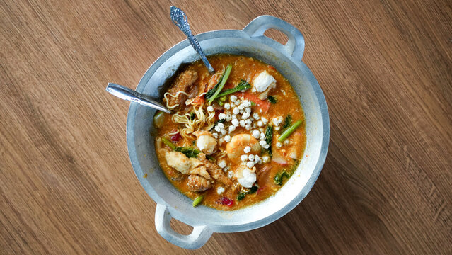 Top view of seblak with various ingredients and vegetables in a wok on wooden table with spoon and fork