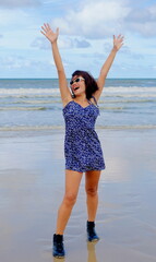 Joyful Asian woman stands confidently on a morning beach, wearing a blue floral dress and black ankle boots, both arms up in the air