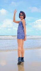 Stylish Asian woman stands confidently on a morning beach, wearing a blue floral dress and black ankle boots, waves back