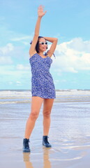 Stylish Asian woman stands confidently on a morning beach, wearing a blue floral dress and black ankle boots, waves with one arm up