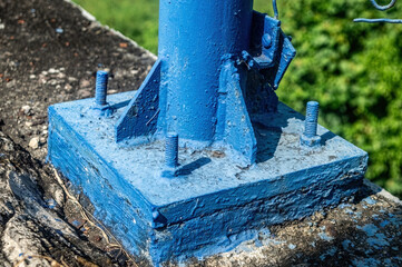 A weathered blue metal base of a pole, secured with bolts on a concrete surface, with a hint of green foliage. industrial resilience, urban infrastructure, and the enduring nature of public works