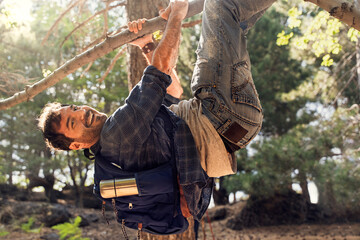 Playful man smiling and hanging upside down from a tree branch while hiking in a forest