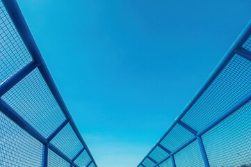 A low angle, symmetrical view of a blue pedestrian bridge with mesh fencing, opening up to a vast,...