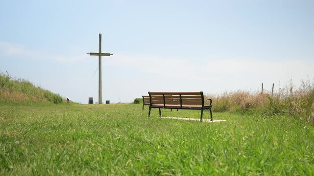 A parch with benches and a large wooden cross in the background. Set against a daytime blue sky.
