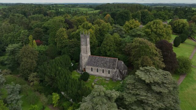 St. Salvator's Church, Glaslough, County Monaghan, Ireland, September 2022. Drone slowly tracking left orbiting around church surrounded by dense trees with tower peering above canopy.