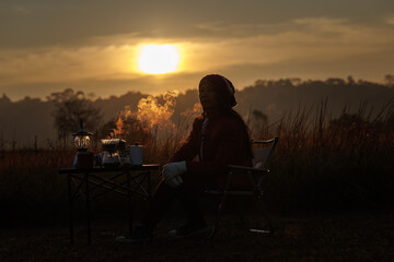 Obraz premium woman sitting outdoors with camping gear, including a table, chair, and a steam lamp. She is enjoying her morning coffee in a serene, natural setting.