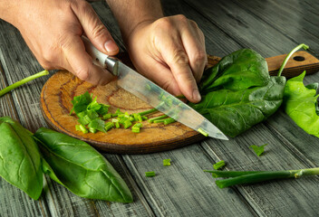 Two hands are skillfully chopping green onions and leafy greens on a wooden cutting board in a...