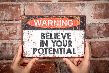 Motivational Warning Sign Saying Believe in Your Potential Held by Hands Against Brick Wall