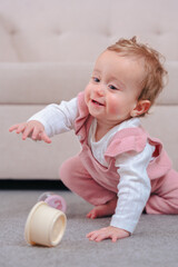 Baby daughter in pink overalls kneels on carpet by beige sofa,reaching to grasp a rolling wooden...