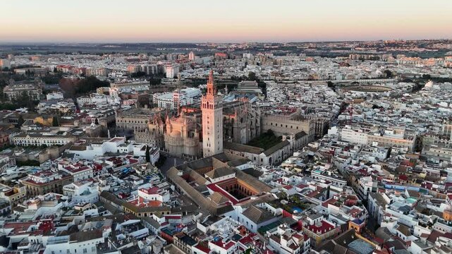 Aerial view of a cityscape at dusk, showcasing a blend of historical and modern architecture. The skyline features prominent towers and rooftops, with a soft pastel sky.