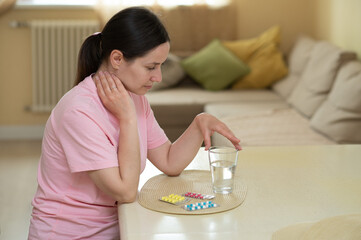 woman sitting at a table, holding her neck in pain and looking at a table filled with colorful pills
