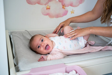 A smiling baby lies on a changing table in a nursery, surrounded by soft pastel decor. A caregiver...