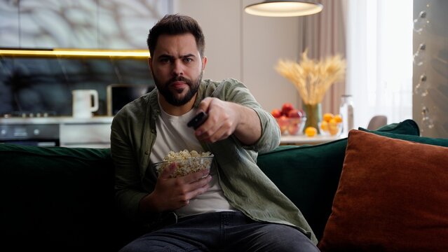 Stunned interested Caucasian man male guy homeowner eating popcorn looking at camera watching TV series film movie media watch television addiction broadcasting using controller indoor home relax room
