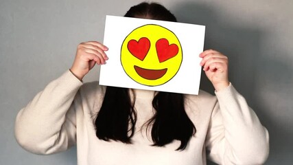 Woman holding heart eyed emoji mask in front of her face. Social media trends, Gen Z marketing, World emoji day 17 July. - Powered by Adobe