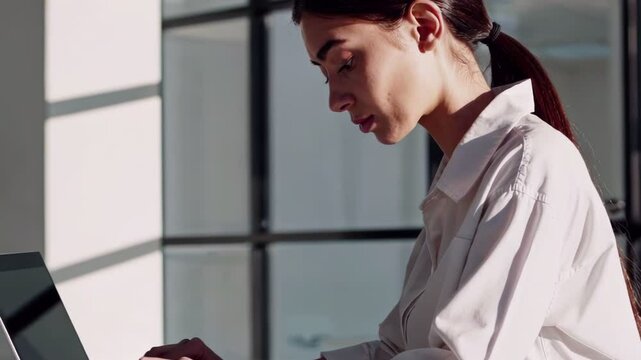 Woman working on laptop in sunlit office, wearing white shirt, side view, focus on face and posture