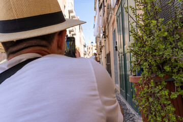Ein Tourist im weißen Hemd und Hut macht Foto mit dem Handy von einer schönen Gasse am Sommertag....
