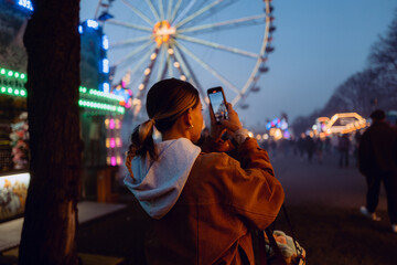 Young woman taking photo of festive fair with Ferris wheel in background, glowing lights and evening atmosphere.
