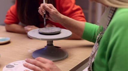 Skilled pottery instructor guiding student while hand painting delicate ceramic plate on rotating support in creative ceramic studio workshop