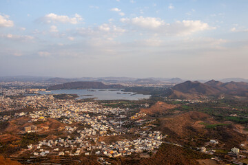 Panoramic aerial view of Udaipur city nestled around its beautiful lakes, surrounded by the Aravalli hills under a vast sky, showcasing Rajasthan's unique urban and natural landscape