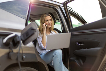Businesswoman working on laptop and talking on phone while charging electric car
