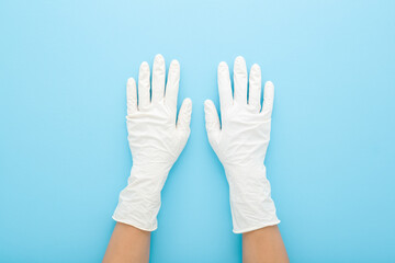 Young adult woman hands in white latex protective gloves on light blue table background. Pastel...