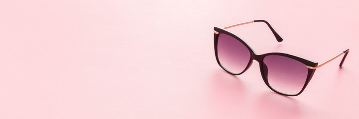 Female sunglasses with dark black plastic frame on light pink table background. Pastel color. Closeup. Wide banner. Empty place for text.